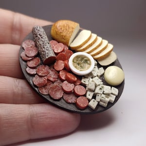 Puede incluir: Una tabla de embutidos en miniatura con salami, pan en rodajas, queso y un pequeño cuenco con salsa. La tabla es de color gris oscuro y los alimentos están dispuestos de forma atractiva. También hay una pequeña bola de queso amarilla.