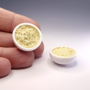 Puede incluir: Dos cuencos blancos en miniatura rellenos de una sustancia texturizada de color amarillo pálido que recuerda a mantequilla con hierbas o salsa. Los cuencos tienen un exterior liso y redondeado y una superficie interior cremosa con remolinos.