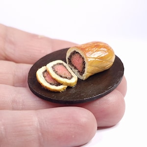 May include: Miniature Beef Wellington on a small, dark brown plate. The pastry is golden brown, with visible layers and a textured surface. Two slices of the wellington are visible, revealing the pink meat and dark filling.