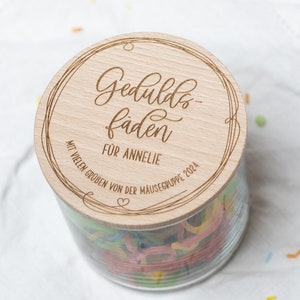 Könnte beinhalten: Ein Holzdeckel mit dem Text "Geduld-fäden für Annelie mit vielen Grüßen von der Mäusegruppe 2024" eingraviert. Der Deckel befindet sich auf einem Glasgefäß, das mit bunten Fäden gefüllt ist.