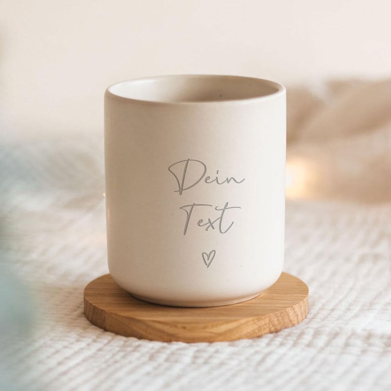 May include: A white ceramic mug with a wooden coaster. The mug has the text "Dein Text" and a small heart printed on it.