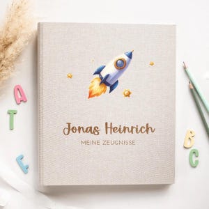 Könnte beinhalten: Ein beigefarbenes Album mit Leinenstruktur und einer Raketenillustration in Blau, Weiß und Orange. Die Rakete fliegt durch den Weltraum mit gelben Sternen. Der Text "Jonas Heinrich" und "Meine Zeugnisse" ist in Braun auf dem vorderen Umschlag gedruckt.