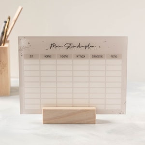 May include: A white acrylic weekly schedule board with the text "Mein Stundenplan" and days of the week labeled in German: Montag, Dienstag, Mittwoch, Donnerstag, Freitag. The board is on a wooden stand.