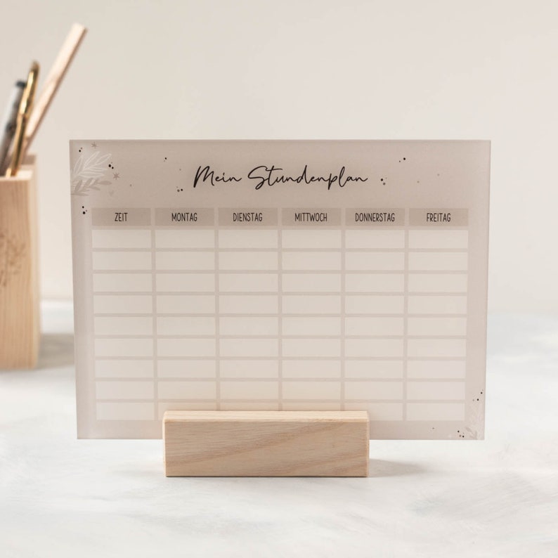 May include: A white acrylic weekly schedule board with the text "Mein Stundenplan" and days of the week labeled in German: Montag, Dienstag, Mittwoch, Donnerstag, Freitag. The board is on a wooden stand.