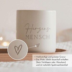 May include: White ceramic mug with the text "Herzeng Mensch" and a small heart design. The mug is on a wooden surface.