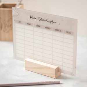 May include: A white acrylic dry erase board with a light brown wood stand. The board has a grid pattern and the text "Mein Stundenplan" at the top. The board is used for scheduling.