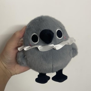 May include: A gray plush bird toy with black eyes, a black beak, and a white ruffled collar. The bird has black feet and is standing on a white surface.