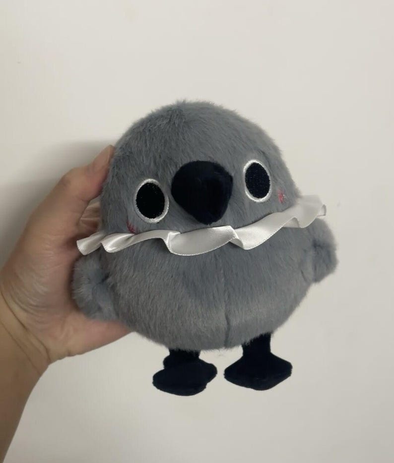 May include: A gray plush bird toy with black eyes, a black beak, and a white ruffled collar. The bird has black feet and is standing on a white surface.