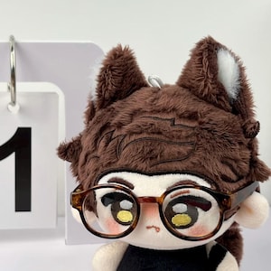 May include: A plush doll with brown fur, pointed ears with white accents, and brown hair. The doll features embroidered eyes with yellow highlights, a small mouth, and wears round, tortoise-shell patterned glasses. A white calendar card with the number 1 is visible.