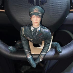 May include: A plush doll in a military uniform, including a hat, jacket, and tie, is positioned on a car steering wheel. The doll's arms and legs are draped over the wheel, creating a unique decorative accent.