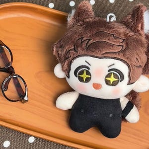 May include: A plush doll with brown hair, pointed ears, and embroidered eyes featuring yellow star-shaped pupils. The doll has a light skin tone, rosy cheeks, and wears a black outfit. Beside the doll are dark-rimmed glasses with brown frames.