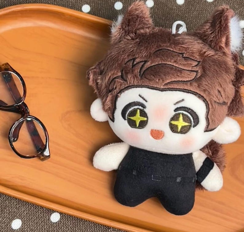 May include: A plush doll with brown hair, pointed ears, and embroidered eyes featuring yellow star-shaped pupils. The doll has a light skin tone, rosy cheeks, and wears a black outfit. Beside the doll are dark-rimmed glasses with brown frames.