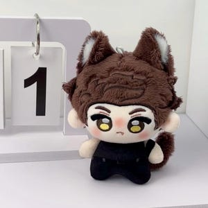 May include: A plush doll featuring brown textured hair, pointed animal ears with white inner details, and large yellow eyes. The doll has a small mouth, rosy cheeks, and wears a black outfit. A metal loop is attached to the top of the head.