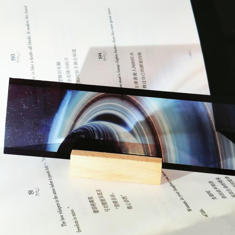 K&ouml;nnte beinhalten: Rechteckiger Lesezeichen mit kosmischem Design, das ein Schwarzes Loch darstellt. Es wird von einem kleinen, rechteckigen Holzblock getragen, der auf einem Buch mit Text liegt.