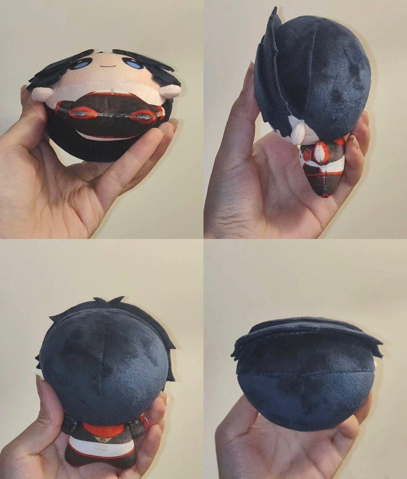 Pu&ograve; includere: Una bambola di peluche con un corpo rotondo e un design di personaggio. La bambola ha capelli neri, carnagione chiara e indossa un abito rosso e bianco. La bambola &egrave; tenuta in mano, mostrando i suoi dettagli da pi&ugrave; angolazioni.
