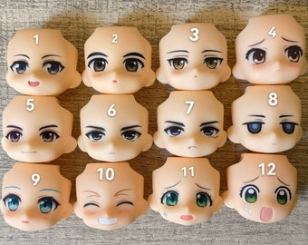 May include: Twelve different blank doll heads with painted facial expressions. The heads are made of a light beige plastic and have different eye colors, hair colors, and facial expressions. The heads are numbered 1 through 12.