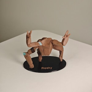 May include: A brown crab figurine with articulated limbs and a black base. The figurine has light blue accents and the name "Rocky" printed on the base. The crab is posed with its claws raised.