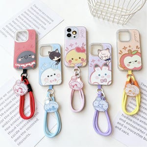 May include: A collection of colorful phone cases with cartoon character designs. Each case has a matching wrist strap. Designs include a bird, cat, chick, bunny, and apple. The cases are on a white surface.
