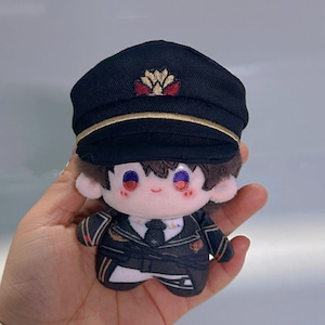 Op de afbeelding: Een pluche speelgoed met bruin haar, dat een zwarte pet draagt met een gouden rand en een rood en gouden embleem. Het speelgoed draagt een zwart-wit uniform met een witte kraag en een zwarte stropdas. Het speelgoed heeft rode wangen en blauwe ogen.