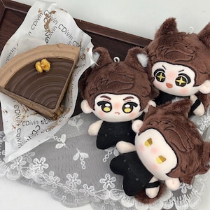 May include: A slice of chocolate cake with a walnut topping on C'Divin branded paper, accompanied by three plush dolls with brown fur, pointed ears, and black outfits. The dolls feature embroidered facial details and star-shaped eyes.