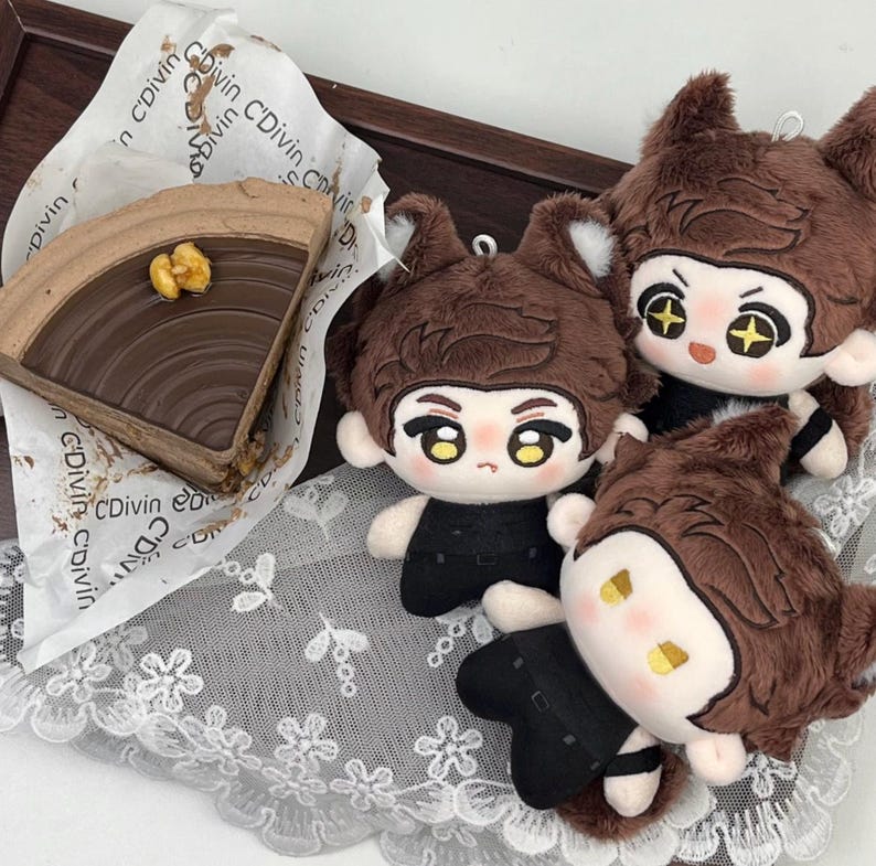 May include: A slice of chocolate cake with a walnut topping on C'Divin branded paper, accompanied by three plush dolls with brown fur, pointed ears, and black outfits. The dolls feature embroidered facial details and star-shaped eyes.