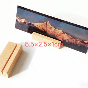 K&ouml;nnte beinhalten: Ein Holz-Fotohalter mit einem Schlitz, der einen Landschaftsdruck eines Bergmassivs h&auml;lt. Der Halter ist 14 cm lang, 6.4 cm breit und 2.5 cm hoch. Der Druck zeigt eine Berglandschaft mit blauem Himmel.