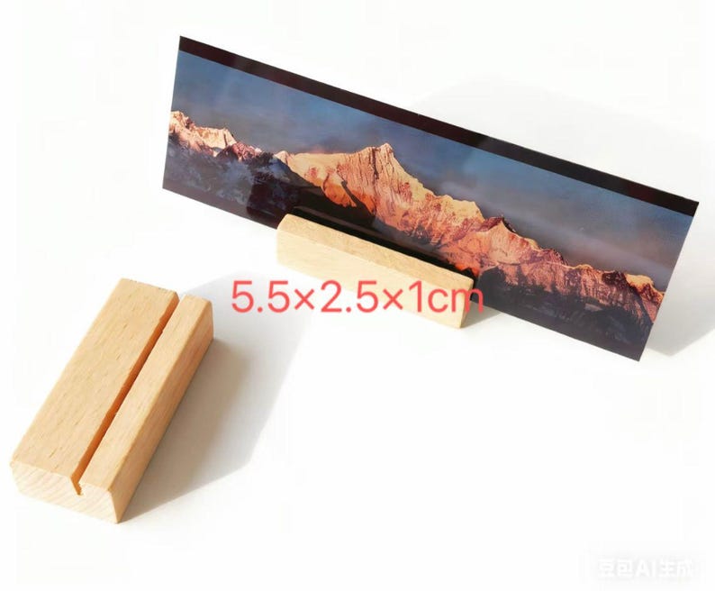 K&ouml;nnte beinhalten: Ein Holz-Fotohalter mit einem Schlitz, der einen Landschaftsdruck eines Bergmassivs h&auml;lt. Der Halter ist 14 cm lang, 6.4 cm breit und 2.5 cm hoch. Der Druck zeigt eine Berglandschaft mit blauem Himmel.