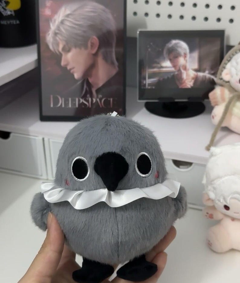 May include: A gray plush bird with a white ruffle collar and black eyes. The bird has a black beak and is standing on a white surface.