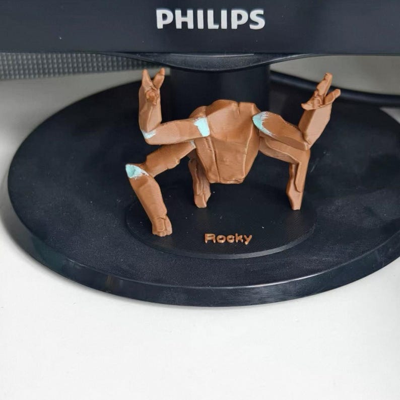 May include: A brown figurine of a creature with outstretched arms and legs, with light blue accents. The figurine is on a black base with the name "Rocky" in small letters. The background includes a computer monitor.