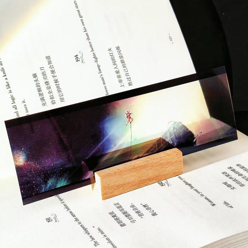 K&ouml;nnte beinhalten: Rechteckiger Kunstdruck mit einer Person auf einem Seil vor einem farbenfrohen Weltraumhintergrund. Der Druck wird auf einem kleinen Holzblock aus Naturholz pr&auml;sentiert, der auf einem offenen Buch liegt. Das Bild hat eine vertr&auml;umte Qualit&auml;t.