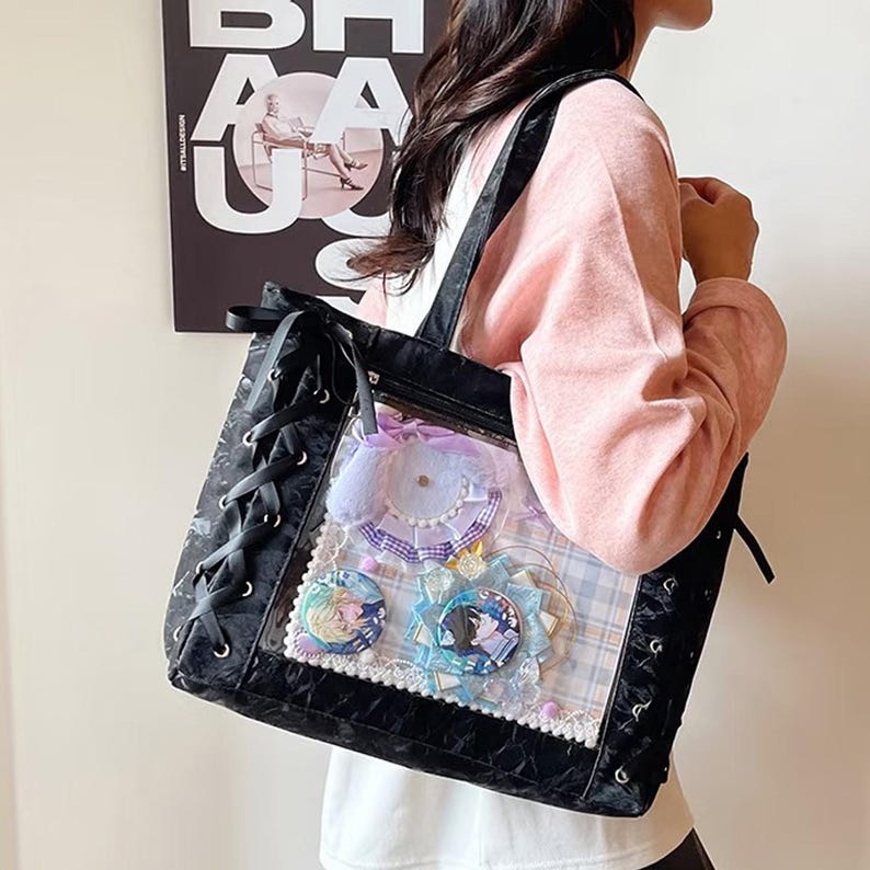 Pode incluir: Bolsa tote preta com um painel frontal transparente exibindo pinos e acess&oacute;rios decorativos. A bolsa tem um design com cadar&ccedil;os nas laterais e al&ccedil;as pretas. O interior apresenta um padr&atilde;o xadrez.