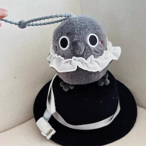 May include: A gray plush owl with a white lace collar, attached to a black hat with a white ribbon. The owl is attached to a gray and white lanyard with a clip.