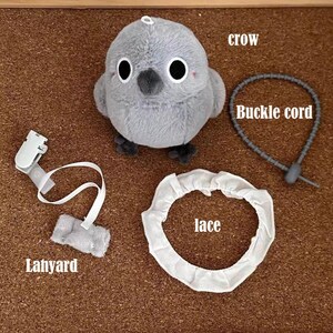 May include: A gray plush crow toy with black eyes and a black beak. The crow is attached to a white lace trim circle and a white lanyard with a plastic clip. The crow also has a gray buckle cord attached to it.