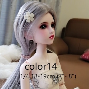 Puede incluir: Una mu&ntilde;eca a escala 1/4 con cabello largo gris plateado peinado en una coleta. La mu&ntilde;eca lleva un vestido blanco sin tirantes con detalles de purpurina plateada. La mu&ntilde;eca tiene una pinza para el cabello con una flor blanca en el cabello. Los ojos de la mu&ntilde;eca son marrones y tiene un ligero rubor rosa en las mejillas. La mu&ntilde;eca mide 18-19 cm (7" - 8"). El texto "color14" es visible en el pecho de la mu&ntilde;eca.