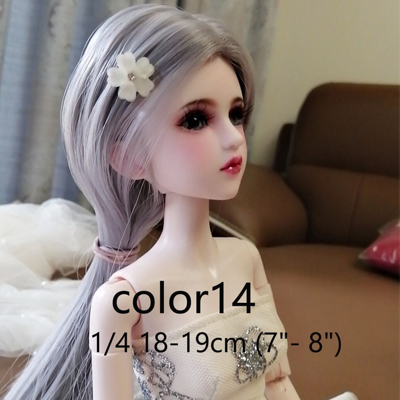 Puede incluir: Una mu&ntilde;eca a escala 1/4 con cabello largo gris plateado peinado en una coleta. La mu&ntilde;eca lleva un vestido blanco sin tirantes con detalles de purpurina plateada. La mu&ntilde;eca tiene una pinza para el cabello con una flor blanca en el cabello. Los ojos de la mu&ntilde;eca son marrones y tiene un ligero rubor rosa en las mejillas. La mu&ntilde;eca mide 18-19 cm (7" - 8"). El texto "color14" es visible en el pecho de la mu&ntilde;eca.