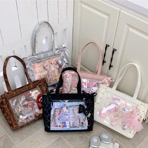 Pode incluir: Cinco sacolas tote em v&aacute;rias cores: marrom, prata, rosa, preto e branco. Cada sacola tem um painel frontal transparente exibindo itens decorativos e imagens. A sacola preta apresenta o texto "Happy Birthday". As sacolas t&ecirc;m diferentes estilos de al&ccedil;a e elementos decorativos.