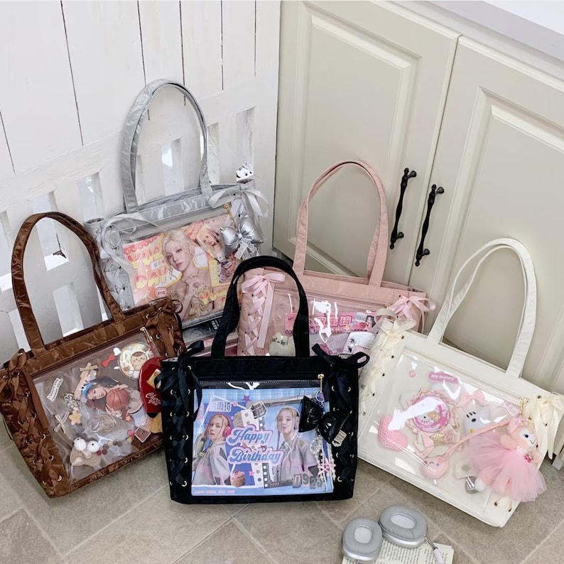 Pode incluir: Cinco sacolas tote em v&aacute;rias cores: marrom, prata, rosa, preto e branco. Cada sacola tem um painel frontal transparente exibindo itens decorativos e imagens. A sacola preta apresenta o texto "Happy Birthday". As sacolas t&ecirc;m diferentes estilos de al&ccedil;a e elementos decorativos.