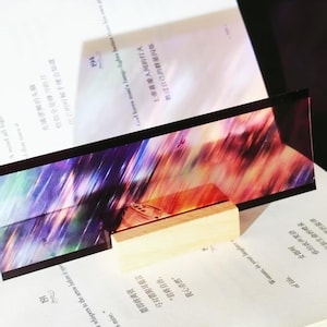 K&ouml;nnte beinhalten: Ein rechteckiger Lesezeichen mit einem farbenfrohen abstrakten Design, das von einem kleinen Holzblock getragen wird, liegt auf einem Buch. Das Lesezeichen weist eine Mischung aus lila, roten und orangen Farbt&ouml;nen auf. Die Buchseiten zeigen gedruckten Text.