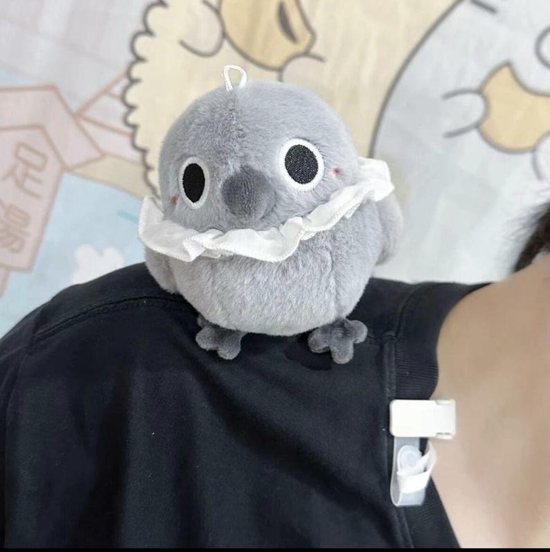 May include: A gray plush bird with a white ruffle around its neck. The bird has large black eyes and a small black beak.