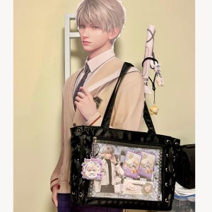 Pode incluir: Bolsa tote preta com um bolso frontal transparente exibindo cart&otilde;es de personagens e um pingente. A bolsa tem al&ccedil;as pretas e costuras decorativas. Uma imagem de papel&atilde;o de uma pessoa em uniforme escolar est&aacute; atr&aacute;s da bolsa.