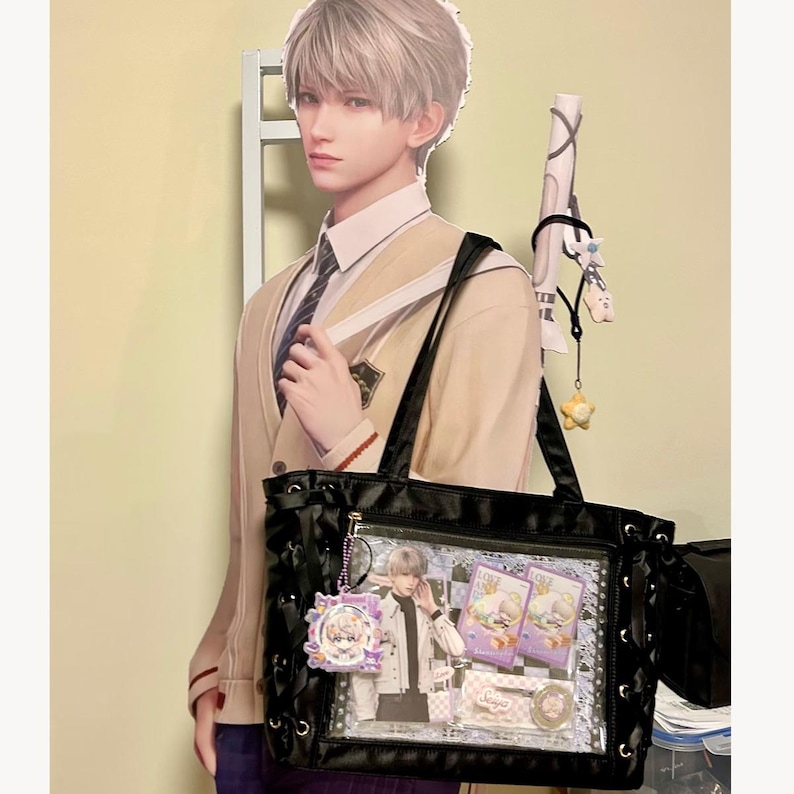 Pode incluir: Bolsa tote preta com um bolso frontal transparente exibindo cart&otilde;es de personagens e um pingente. A bolsa tem al&ccedil;as pretas e costuras decorativas. Uma imagem de papel&atilde;o de uma pessoa em uniforme escolar est&aacute; atr&aacute;s da bolsa.