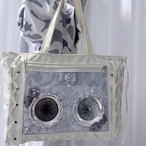 Pode incluir: Uma mala tote branca com um painel frontal decorativo cinza e prateado. A mala apresenta um fecho de correr, detalhes de atacadores nas laterais e dois elementos decorativos circulares. A mala tem al&ccedil;as brancas e um la&ccedil;o com uma joia.
