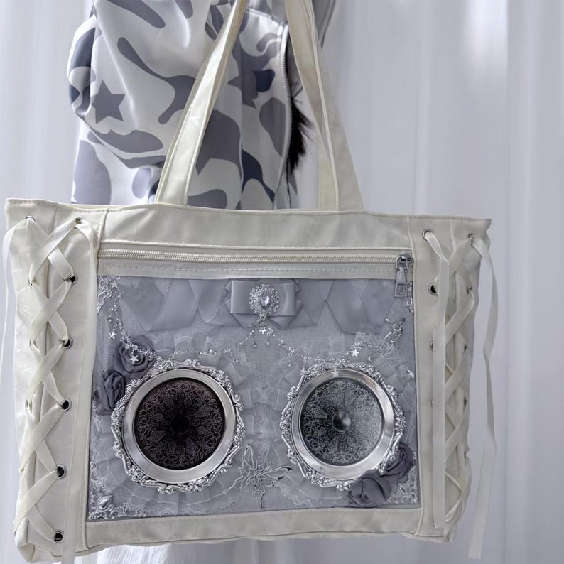 Pode incluir: Uma mala tote branca com um painel frontal decorativo cinza e prateado. A mala apresenta um fecho de correr, detalhes de atacadores nas laterais e dois elementos decorativos circulares. A mala tem al&ccedil;as brancas e um la&ccedil;o com uma joia.