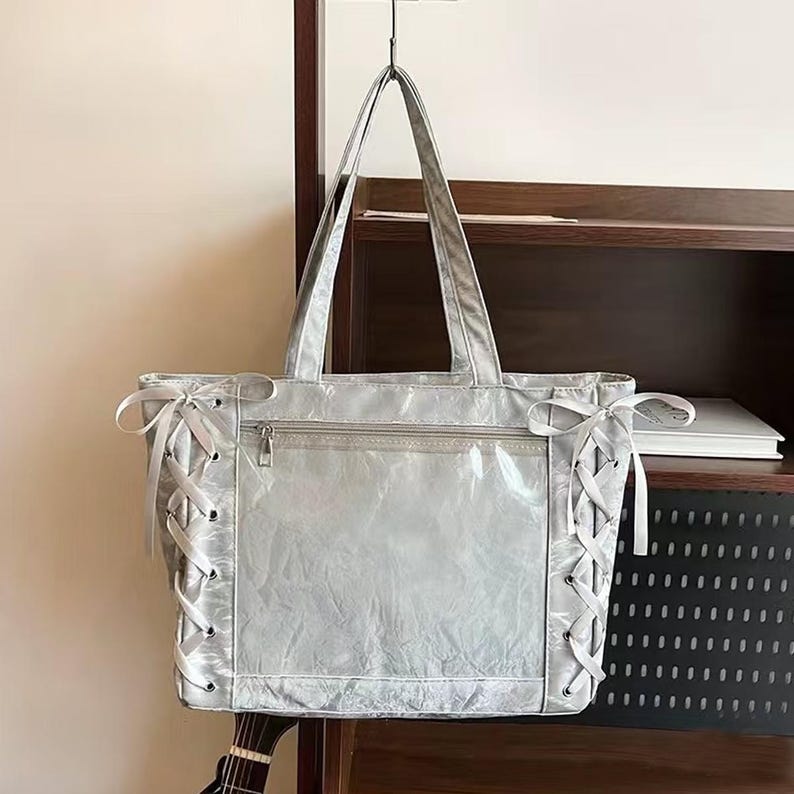 Pode incluir: Uma bolsa tote prateada com fecho de correr e al&ccedil;as compridas. Os lados apresentam um design com atacadores e fitas brancas. A bolsa est&aacute; pendurada num gancho preto.