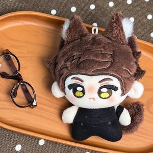 May include: A plush doll with brown fur, pointed ears, and a white face featuring large yellow eyes and a small mouth. The doll wears a black outfit and has a small metal loop on top of its head. Beside the doll are round brown-rimmed glasses.