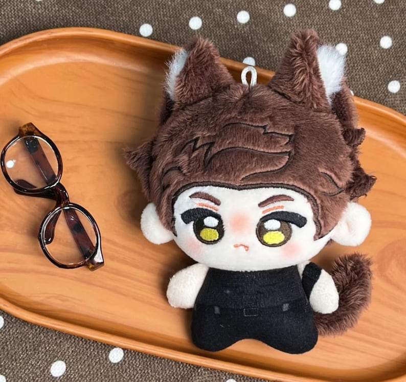 May include: A plush doll with brown fur, pointed ears, and a white face featuring large yellow eyes and a small mouth. The doll wears a black outfit and has a small metal loop on top of its head. Beside the doll are round brown-rimmed glasses.