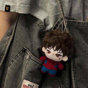 May include: A small plush doll with brown curly hair and a red and blue spider-themed outfit, attached to a silver ball chain. The doll is tucked into the pocket of a dark gray denim garment with a Lee brand label visible on the lower pocket area.