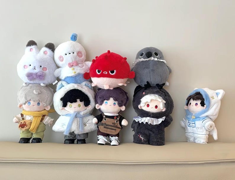 K&ouml;nnte beinhalten: Eine Sammlung von Pl&uuml;schpuppen in verschiedenen Designs, darunter ein Hase, ein Vogel und mehrere Puppen in Outfits. Die Puppen sind in verschiedenen Farben gehalten, darunter Wei&szlig;, Rot, Grau und Schwarz. Sie sind auf einer beigen Oberfl&auml;che angeordnet.