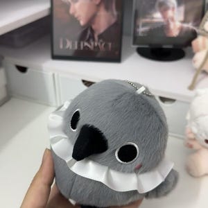 May include: A gray plush bird with a white ruffle collar. The bird has black eyes and a black beak. The bird is a keychain.
