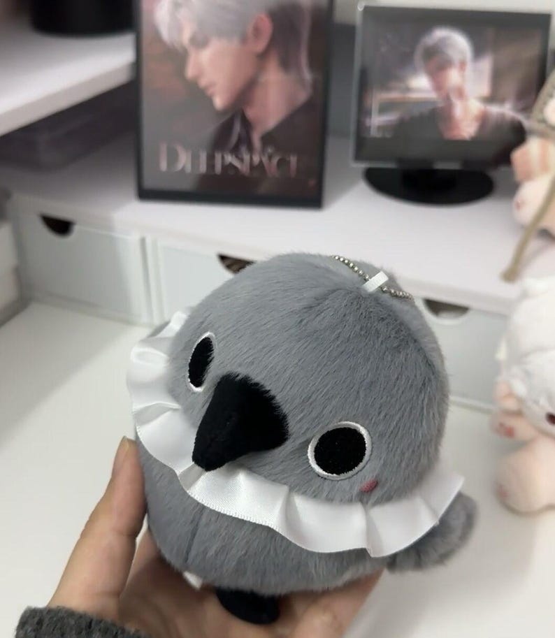 May include: A gray plush bird with a white ruffle collar. The bird has black eyes and a black beak. The bird is a keychain.